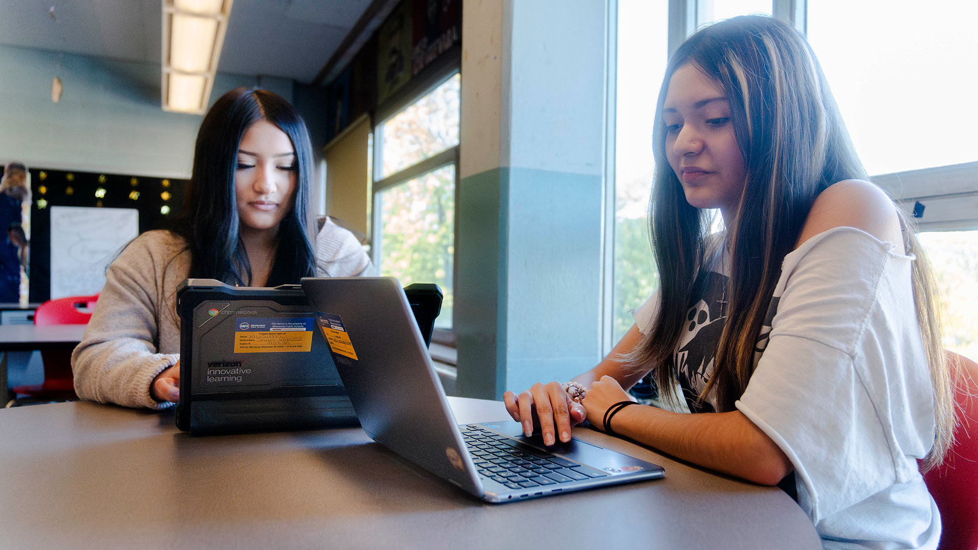 Two students sit by a classroom window and use laptops.
