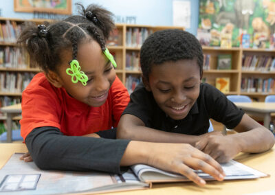 Two young students at a table in a library, smile as they look at a picture book.