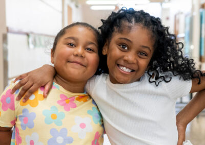 Two young children smile in a hallway with their arms around one another.