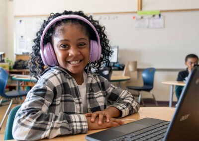 A young child smiles with headphones on as she uses a computer.