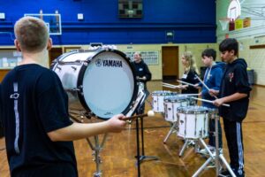 Middle school students practice drumline in a gym, playing bass and snare drums.