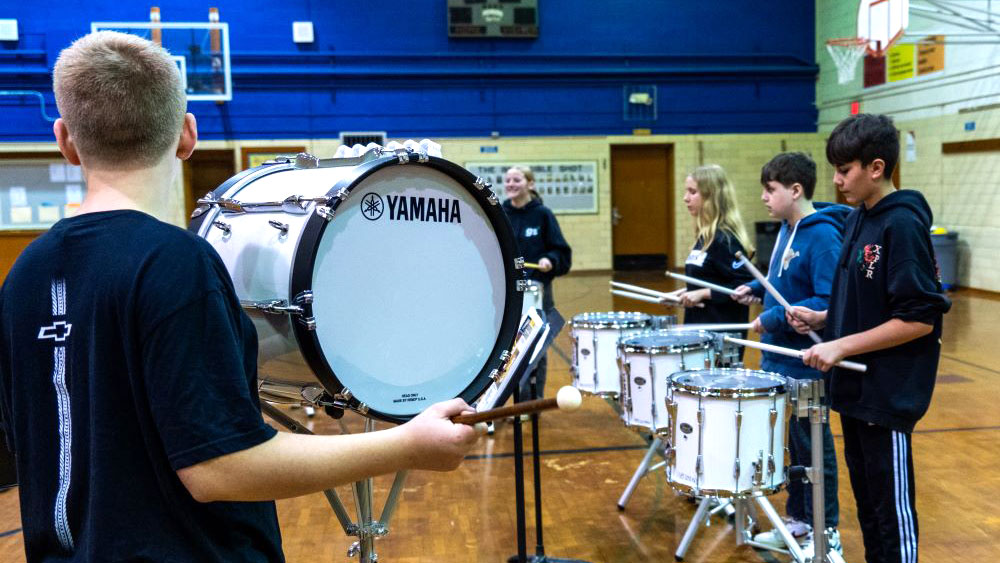 Middle school students practice drumline in a gym, playing bass and snare drums.