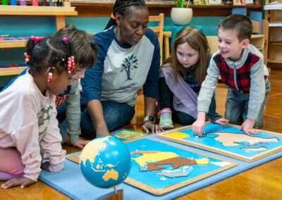 A teacher sits on the floor with her young students as they work on a wooden puzzle of North America.