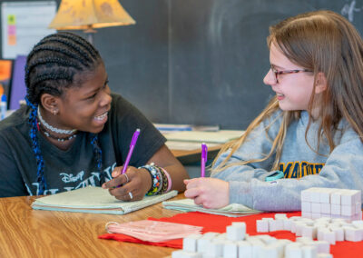 Two girls smile at each other as they write in their notebooks.