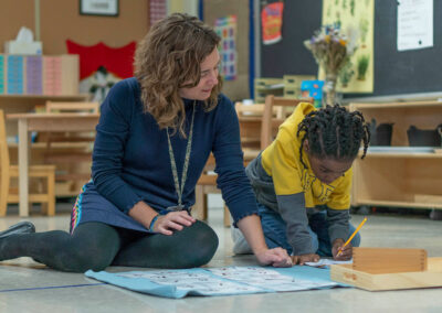 A teacher sits on the floor with her young student as he writes in a notebook.