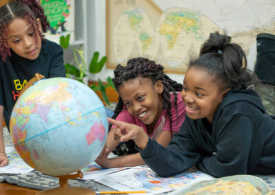 Three girls lay on the floor and point to a globe.