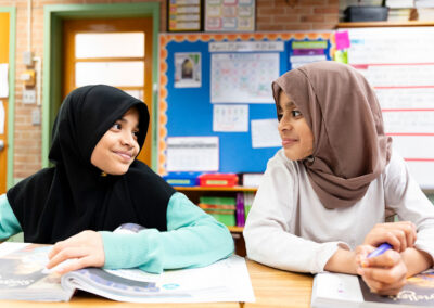 Two girls in a classroom smile at one another.