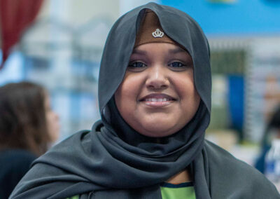 A girl wearing a hijab in a classroom smiling.