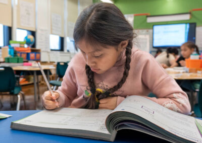 A girl at a table in a classroom writes in a workbook.