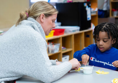 A teacher helps a young student with glue.