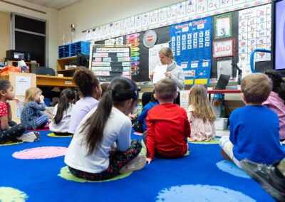 Young children sit on a colorful rug and look up at their teacher.