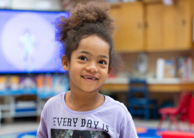 A young child in a classroom missing her front teeth as she smiles.