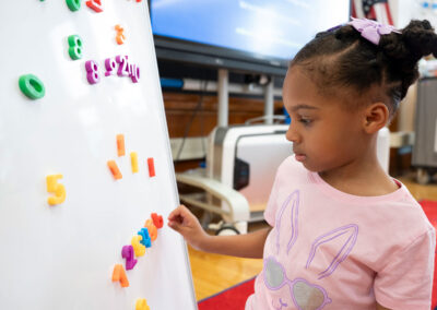 A young child in a classroom adds magnetic numbers to a whiteboard.