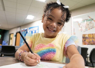 A smiling girl sits at a table with a colored pencil in her hand, ready to draw.