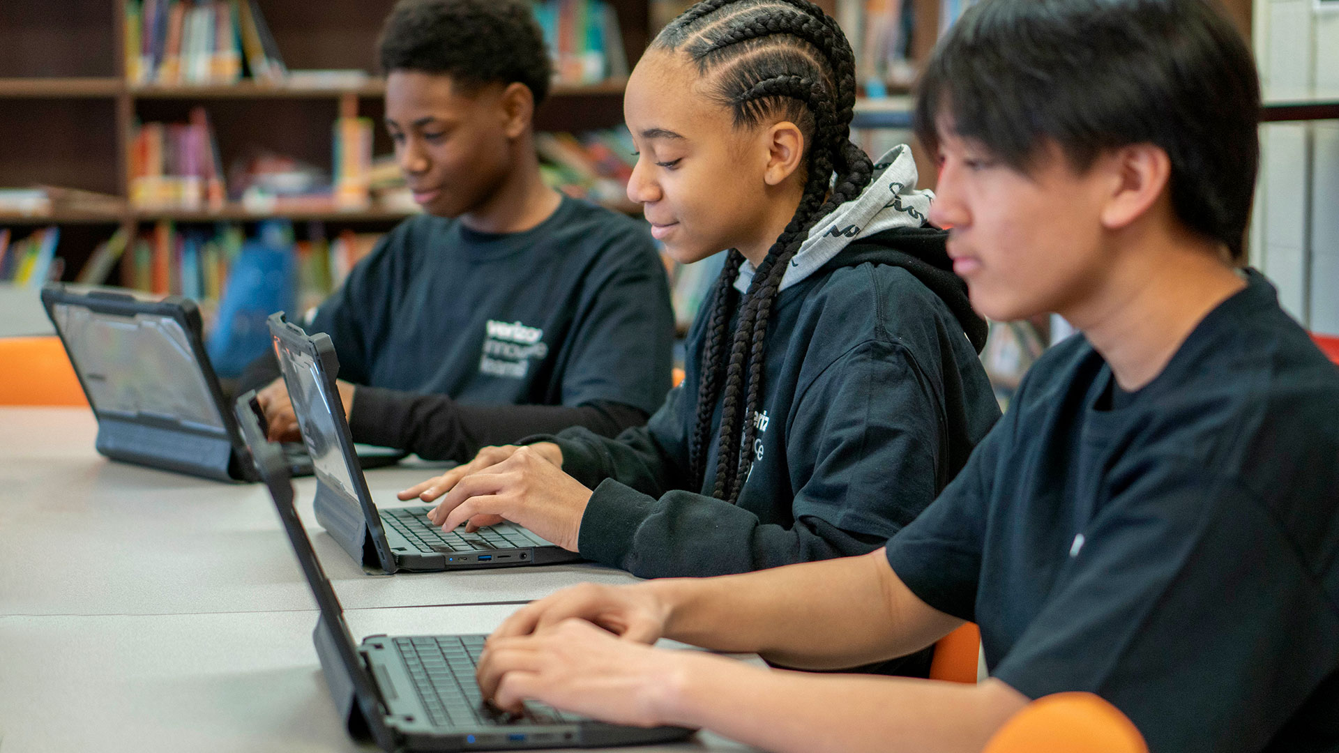 Three students wearing matching shirts sit at a table and type on laptops.