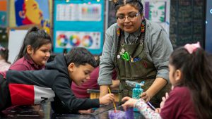 A teacher with an art smock watches as young students paint.