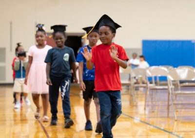 Young children walk in a line wearing graduation caps.