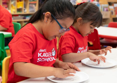 Niños pequeños en un aula practican la escritura de letras con arena en platos de papel.