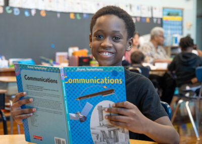 A boy smiles as he holds up a book about communications.
