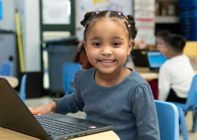 A young girl with a laptop on the table in front of her, smiling.