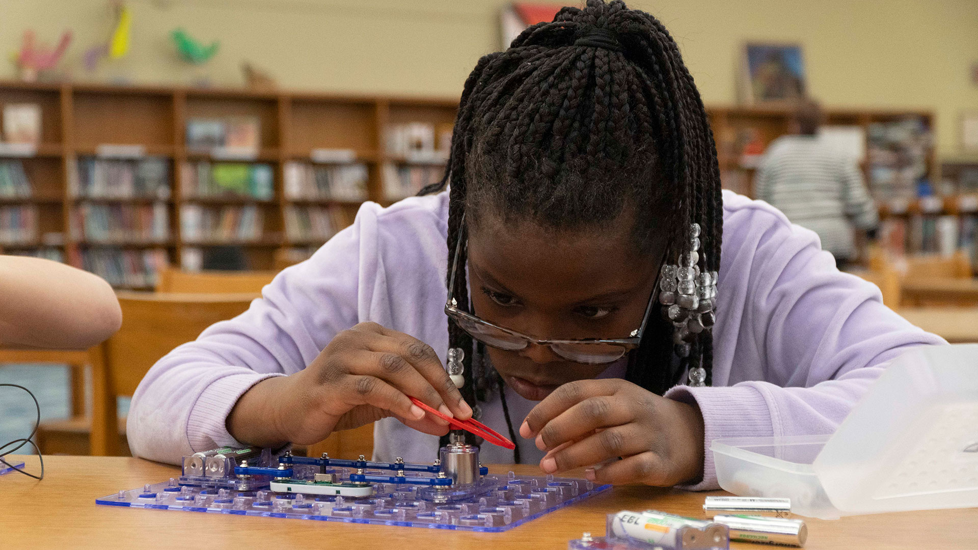 A girl at a table in a library putting an electric device together.