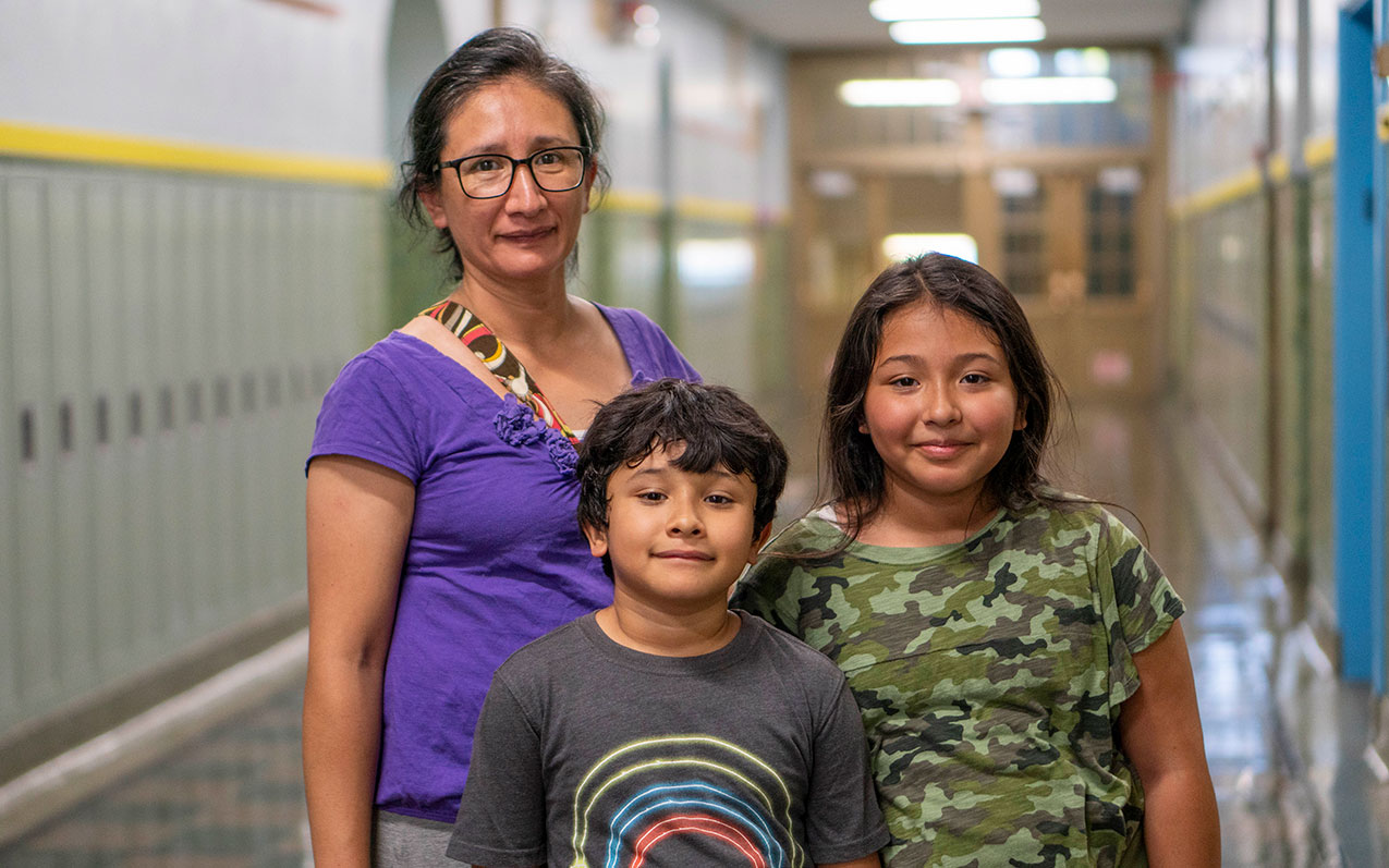 A mother stands with her two students in a hallway with lockers.
