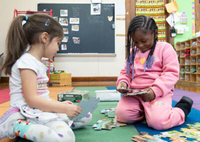 Two young children sit on a colorful rug and put a puzzle together.