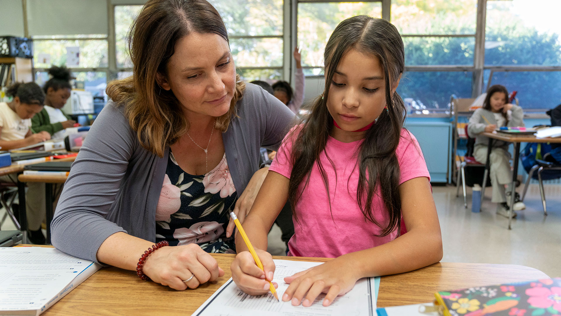 A teacher sits next to her student and helps them with an assignment.