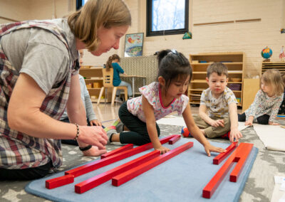 A teacher and her young students on the floor placing long square dowels next to one another.