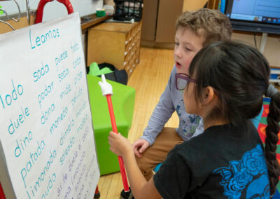 A young girl uses a pointer to point to words on a board while a young boy reads along.