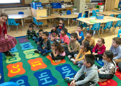 A class of young students sit on a colorful carpet and listen as the teacher reads to them.