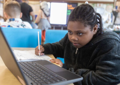 A girl takes notes on paper as she looks at a computer.