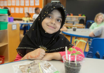 A girl in a hijab smiles with a small plant on the table in front of her.